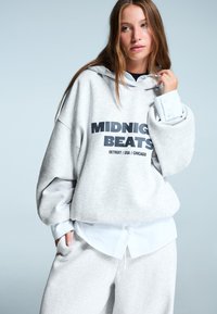 Svetlosivý oversized mikina s kapucňou, na ktorej je nápis "MIDNIGHT BEATS", preložená na bielu košeľu s gombíkmi a doplnená zladenými teplákmi.