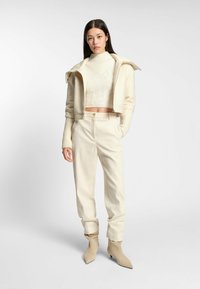 Woman standing wearing cream zip-up jacket, cropped sweater, high-waist pants, and pointed-toe boots against plain white background.