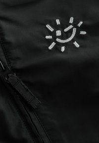 Schwarze Jacke mit glatter Textur, ausgestattet mit einem silbernen gestickten Sonnenmotiv und einem sichtbaren Reißverschluss.