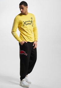Långärmad gul t-shirt med svart grafisk design; parat med svarta sweatpants med röd djurbroderi och vita sneakers.