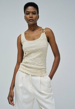 Débardeur beige texturé avec un col arrondi et un matériel doré décoratif sur une épaule. Assorti à un pantalon blanc taille haute.