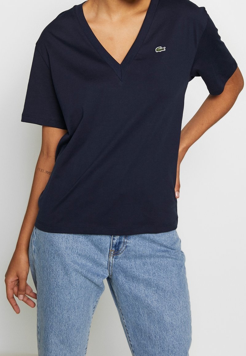 Personne portant un t-shirt bleu marine à col en V avec un petit logo crocodile vert et un jean en denim bleu clair, main sur la hanche.