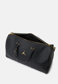 Svart duffelbag laget av teksturert materiale, med gullakenter, en logo, doble håndtak og en glidelåsåpning for enkel tilgang.