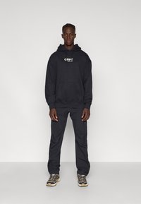 Sudadera negra con bolsillo frontal tipo canguro y logo, combinada con pantalones oscuros y zapatos resistentes en una paleta de colores gris y marrón.