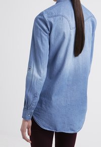 Ljusblå denimskjorta med långa ärmar, rundad kant, knappar vid ärmsluten och en urtvättad effekt. Modell med ryggen mot kameran, med håret nedsläppt.