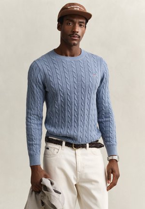 Mann trägt hellblauen Zopfpullover, beige Hose, braunen geflochtenen Gürtel, Uhr, braune Mütze und hält eine beige karierte Jacke in der linken Hand.