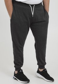 Mann trägt dunkelgraue Sweatpants mit weißem Kordelzug und schwarze Sneakers mit weißen Sohlen und steht mit den Händen in den Taschen.