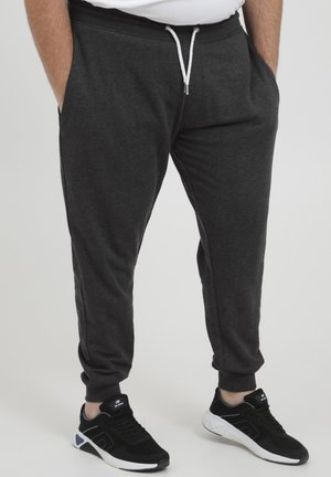 Homme portant un pantalon de survêtement gris foncé avec un cordon blanc et des baskets noires avec des semelles blanches, se tenant les mains dans les poches.