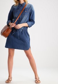 Denim-paitapuku pitkillä hihoilla, napitettu kaulus, nauhavyötärö, sivuhalkiot ja syvän sininen väri; yhdistetty ruskeaan olkalaukkuun.