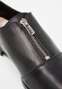 Chaussure en cuir noire avec une fermeture éclair argentée traversant en diagonale la partie avant supérieure, avec des coutures détaillées le long des bords.