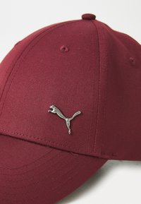 Căciulă maroon dintr-un material texturat, având un logo Puma argintiu lucios pe partea din față. Are detalii cu cusături și o vizieră curbată.