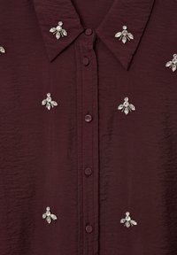 Camicia bordeaux con bottoni realizzata in un tessuto testurizzato, dotata di ornamenti floreali in strass argentati sia sul davanti che sul colletto.