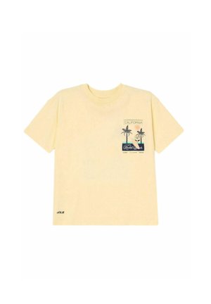 Ung pige står smilende med hænderne i lommerne iført en creme t-shirt med palmeprint, denimshorts og beige slip-on sko.