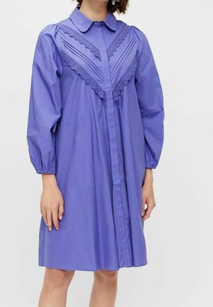 Nainen, joka pukeutunut polvipituiseen, pitkähihainteseen purppuranpunaiseen mekkoon, jossa on terävä kaulus ja koristeellinen pitsireunus V-muodossa rinnassa.