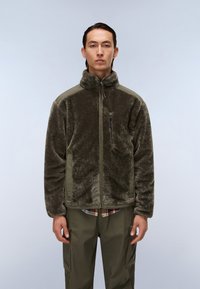 Fleece takki tummanoliivinvärisellä korkealla kauluksella, vetoketjukiinnityksellä ja teksturoidulla kankaalla. Yhdistetty oliivinvärisiin housuihin ruudullisen paidan päällä.
