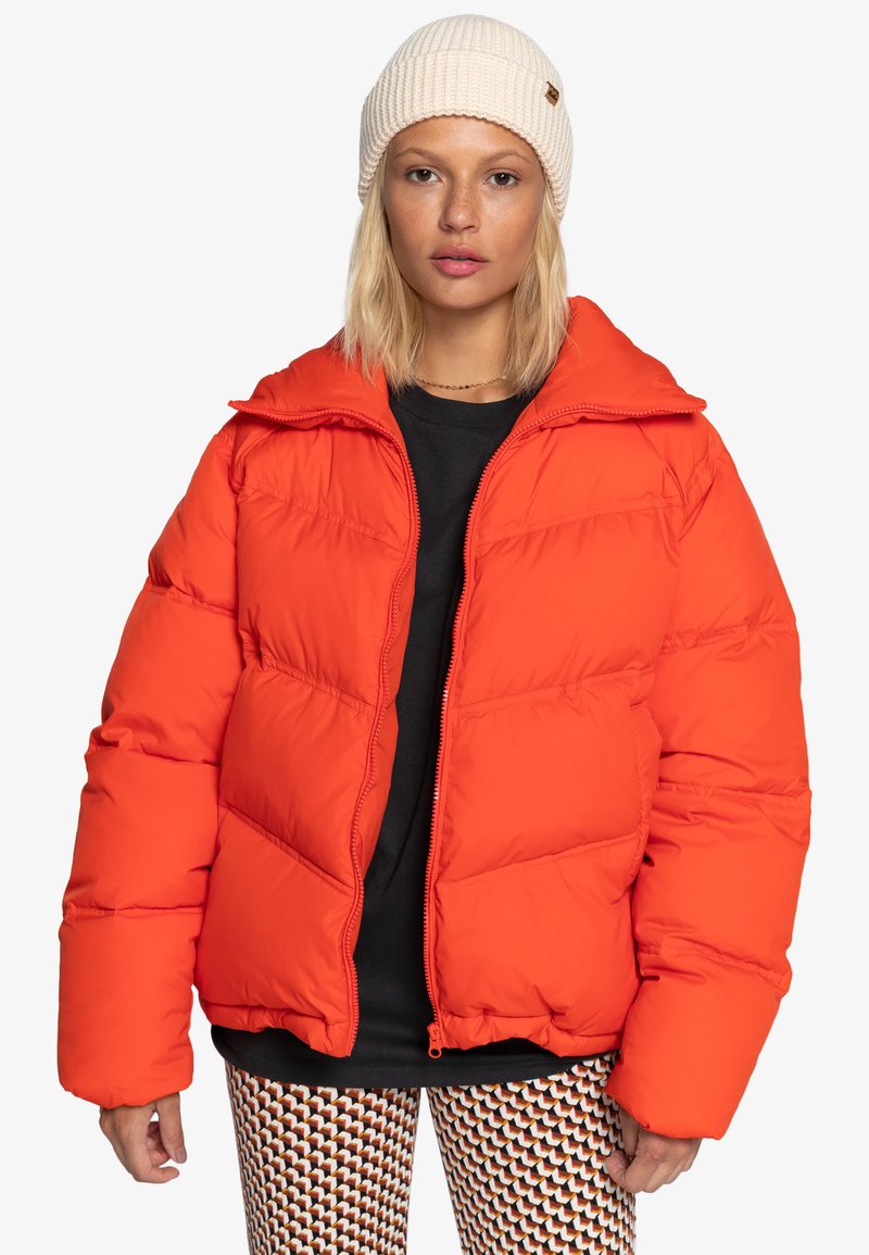 Giacca puffer arancione brillante con zip frontale, vestibilità oversize, scomparti cuciti e colletto alto. Indossata sopra una maglietta nera e pantaloni a fantasia.