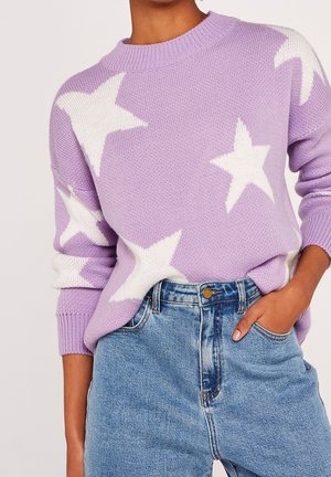 Personne portant un pull en tricot lavande avec de grandes étoiles blanches, associé à un jean en denim bleu taille haute, main dans la poche.
