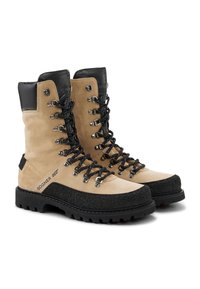 Béžové semišové boty s černými koženými detaily, kovovými očky a černými gumovými podrážkami; navrženy pro odolnost s texturovanými detaily.