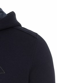 Granatowa bluza z kapturem z teksturowanym kołnierzem, detalem szwu na ramionach oraz geometrycznym logo na klatce piersiowej w jaśniejszym odcieniu szarości.