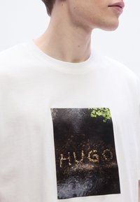 Λευκό βαμβακερό T-shirt με ορθογώνιο γραφικό τύπωμα που απεικονίζει το όνομα "Hugo" φτιαγμένο από πέτρες σε βρεγμένο σκοτεινό φόντο με φυλλώματα.