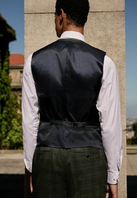 Schwarze Satinweste mit verstellbarem Rückenriemen, getragen über einem weißen Hemd. Grüne karierte Hose sichtbar, mit Knöpfen und Taschen.