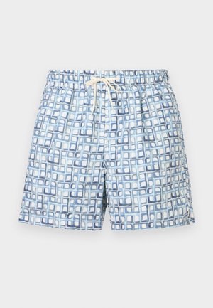 Homme torse nu aux cheveux courts et foncés portant un short de bain bleu et blanc à motifs, debout pieds nus les mains dans les poches sur un fond uni.