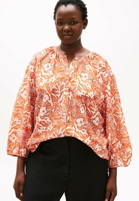 Langærmet bluse i en lys orange og hvid blomsterprint, med en rynket halsudskæring og løs pasform, kombineret med sorte bukser.