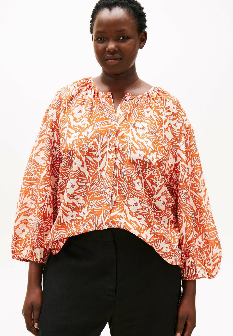Langærmet bluse i en lys orange og hvid blomsterprint, med en rynket halsudskæring og løs pasform, kombineret med sorte bukser.
