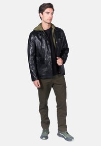 Giacca di pelle nera con chiusura a bottoni e tasca sul petto, sovrapposta a una felpa con cappuccio verde oliva. Abbinata a pantaloni cargo olive e sneakers.