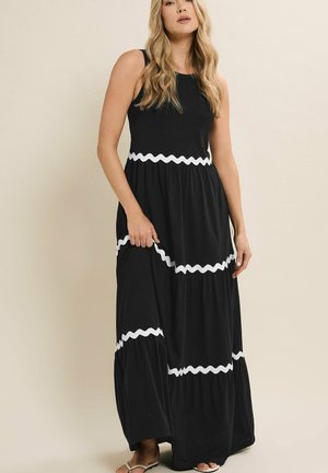 Frau trägt ein ärmelloses schwarzes Maxikleid mit weißen wellenförmigen horizontalen Streifen und beigen Sandalen, steht vor einem neutralen Hintergrund.