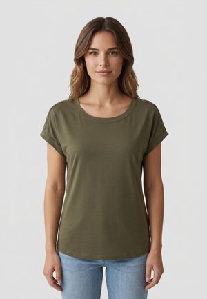 Donna con capelli castani ondulati che indossa una camicia verde oliva a maniche corte con polsini ripiegati e jeans azzurri, in piedi davanti a uno sfondo semplice.