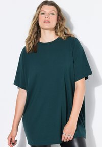 Žena s vlnitými blond vlasy, velkými kruhovými náušnicemi, zeleným očním makeupem, tmavě zeleným oversized tričkem a černými kalhotami na bílém pozadí.