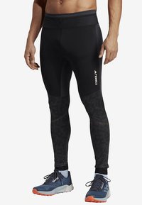 Schwarze sportliche Leggings mit tailliertem Design, strukturierten Einsätzen und einem weißen Logo an der Seite. Kombiniert mit blauen Laufschuhen.