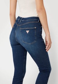 Jeans de mezclilla azul oscuro con una cintura alta, que presentan un parche en forma de triángulo en el bolsillo trasero y detalles de costura sutiles.