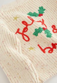 Sweter dzianinowy w kolorze kremowym z kolorowym haftem. Posiada czerwony napis kursywą, zielone liście ostrokrzewu oraz złotą gwiazdkę jako akcent. Teksturowana powierzchnia.