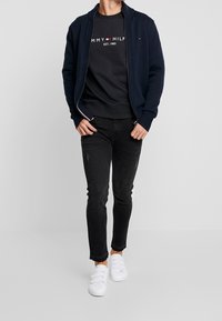 Cárdigan navy con cremallera sobre una camiseta negra con texto blanco, combinado con jeans slim-fit negros y zapatillas blancas sin cordones.