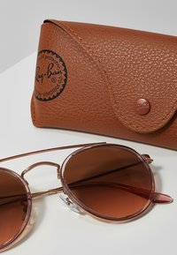 Slnečné okuliare Ray-Ban s okrúhlymi hnedými sklami a tenkými rámami v ružovom zlate, doplnené o hnedú textúrovanú koženú tašku s tlačným gombíkom.