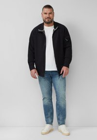 Zwarte jack met rits aan de voorkant en grijze accenten, gecombineerd met een wit T-shirt en lichtblauwe jeans. Afgerond met witte schoenen. Effen achtergrond.
