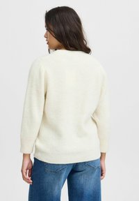Un pull blanc côtelé avec un col rond et des manches trois-quarts, associé à un jean bleu. Texture douce et coupe décontractée.