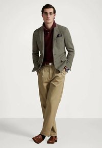 Muž v brýlích, olivovém saku, vínovém polo tričku, béžových kalhotách a hnědých botách, stojící se zkříženýma rukama v kapsách na jednobarevném pozadí.