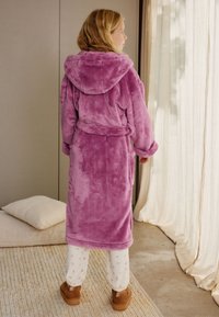Halat de baie din fleece, de culoare violet deschis, cu glugă, talie strânsă și buzunare frontale, purtat peste pantaloni de pijama cu model floral și papuci.