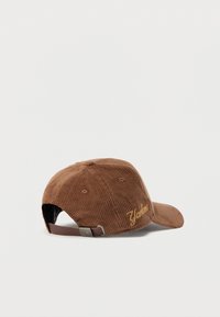 Brown corduroy baseball cap with embroidered yellow text on the side, adjustable strap at the back, and textured ribbed surface.