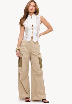 Femme portant un top blanc en dentelle sans manches avec des liens à l'avant et un pantalon cargo beige à jambes larges avec des poches foncées, debout la main sur la hanche.