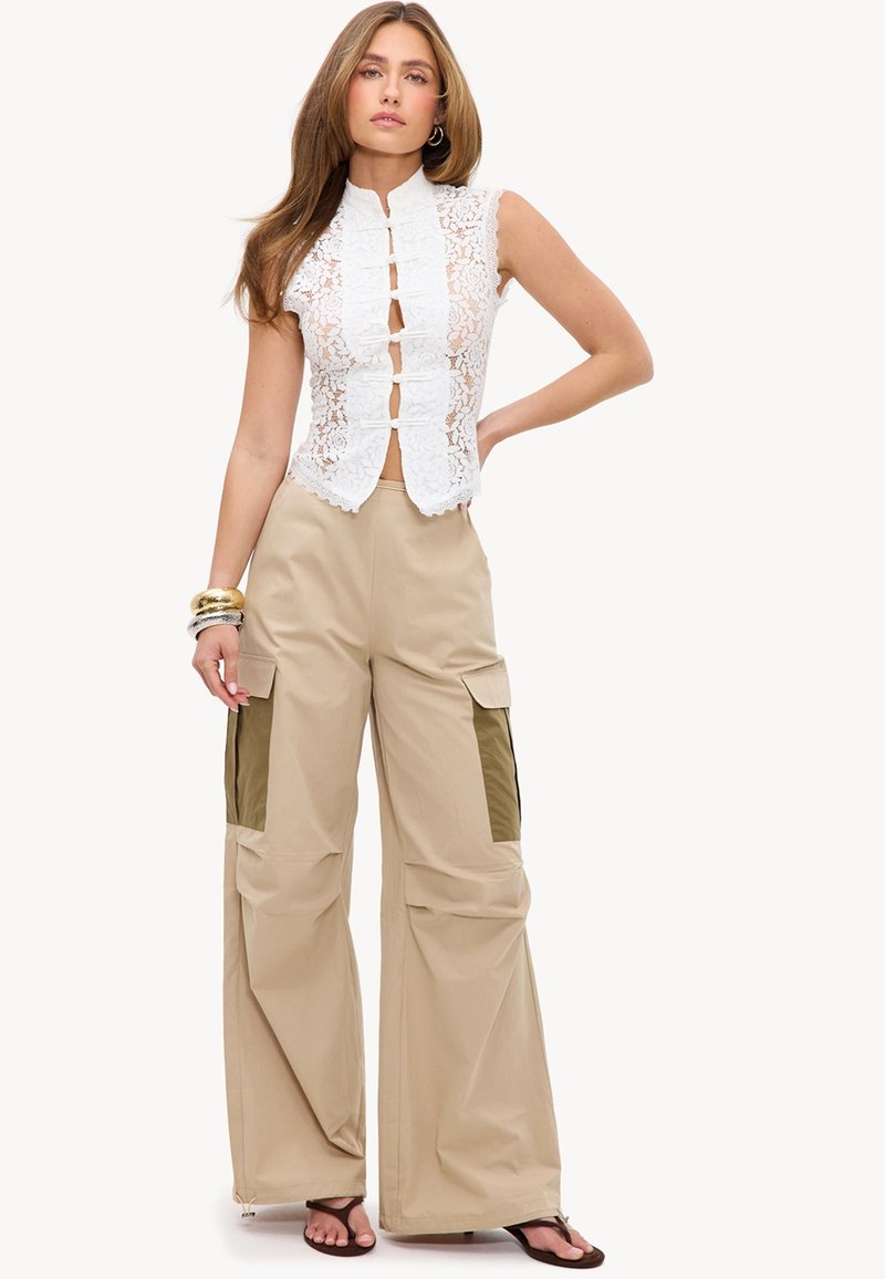 Femme portant un top blanc en dentelle sans manches avec des liens à l'avant et un pantalon cargo beige à jambes larges avec des poches foncées, debout la main sur la hanche.