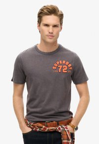 Camiseta de algodón gris con cuello redondo, que presenta el texto en naranja "SUPERDRY 72". Combinada con un cinturón de cuero marrón y una camisa de franela atada a la cintura.