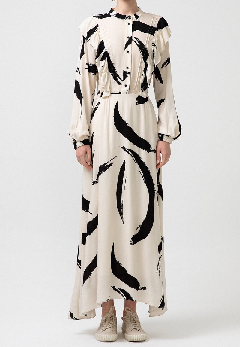 Femme portant une longue robe crème avec des motifs abstraits en coups de pinceau noirs, devant boutonné, manches longues, et des baskets beiges sur fond blanc.
