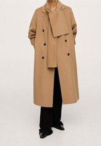 Manteau croisé couleur chamel en laine, avec un col large, des boutons noirs et une écharpe assortie, porté avec un pantalon large noir.