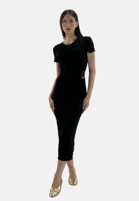 Das Model posiert selbstbewusst in einem figurbetonten schwarzen Kleid mit kurzen Ärmeln, akzentuiert durch eine goldene Schnalle, kombiniert mit glänzenden goldenen Absatzschuhen.