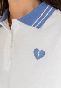 Polo bianco con colletto blu e due strisce bianche. Cuore blu ricamato con simbolo di rottura sul petto.