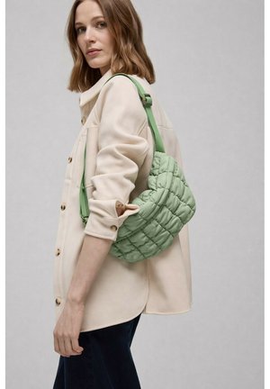 Mujer con cabello castaño claro hasta los hombros, vestida con chaqueta beige abotonada y lleva un bolso de hombro verde claro acolchado sobre fondo gris.
